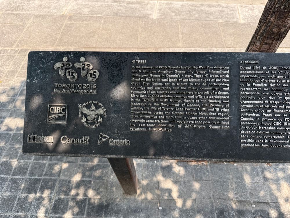 Plaque commemorating the 2015 Pan Am/Parapan Am Games in Toronto, titled “41 Trees.” The plaque features the Toronto 2015 logo, and logos of major partners including CIBC, the Mississaugas of the New Credit First Nation, City of Toronto, Government of Canada, and Government of Ontario.

Text (English side):

⸻

41 TREES

In the summer of 2015, Toronto hosted the XVII Pan American and V Parapan American Games, the largest international multi-sport Games in Canada’s history. These 41 trees, which stand on the traditional lands of the Mississaugas of the New Credit First Nation, are in tribute to the 41 participating countries and territories, and the talent, commitment and teamwork of the athletes who were here in pursuit of a dream.

More than 10,000 athletes, coaches and officials participated in the TORONTO 2015 Games, thanks to the funding and leadership of the Government of Canada, the Province of Ontario, the City of Toronto, Lead Partner CIBC and 15 other municipalities across the Greater Golden Horseshoe region, three universities and more than a dozen other civic-minded corporate sponsors. None of it would have been possible without the passionate dedication of 23,000-plus Games-time volunteers. United We Play.