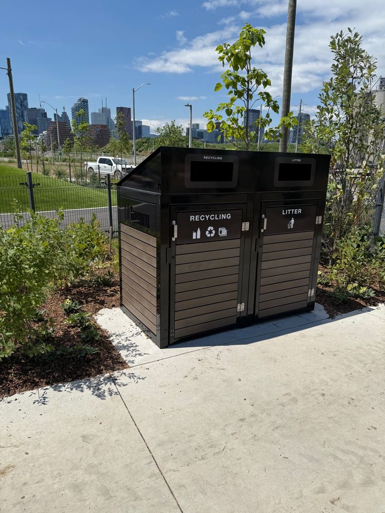 A dual-compartment waste bin for recycling and litter stands on a paved path in Biidaasige Park in Toronto’s Port Lands. The modern black-and-wood bin features the City of Toronto logo. In the background, the downtown skyline rises beyond newly planted greenery and a fenced urban meadow.
