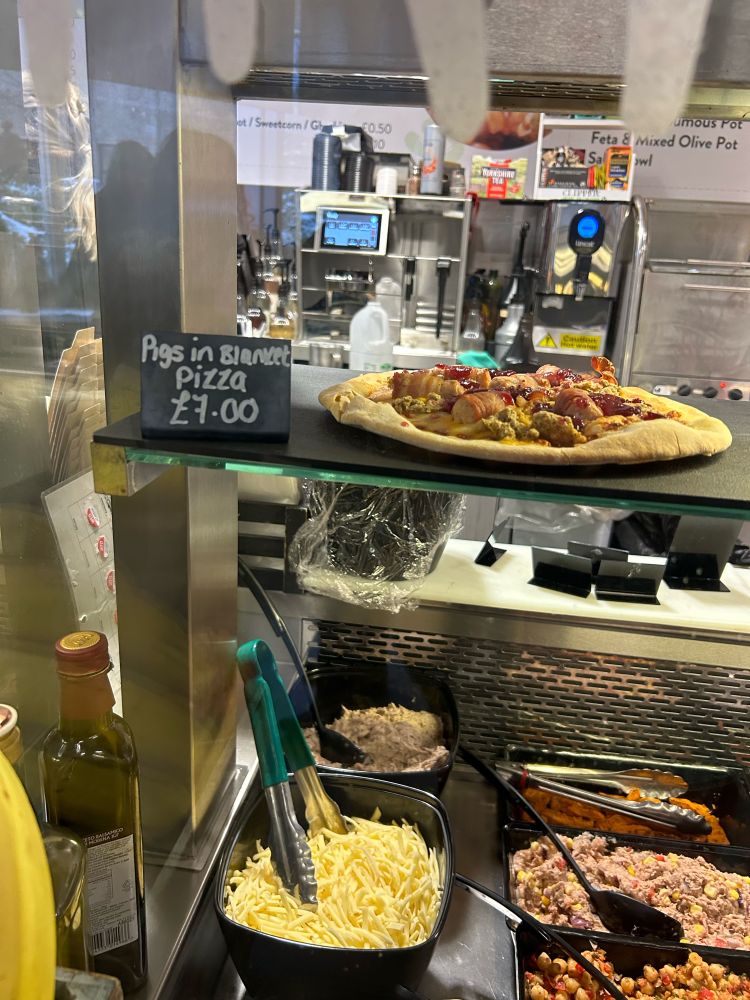 a photo of a pizza topped with pigs-in-blankets, cranberry sauce and stuffing. it’s being sold in my university café, and is shown as costing £7.00.