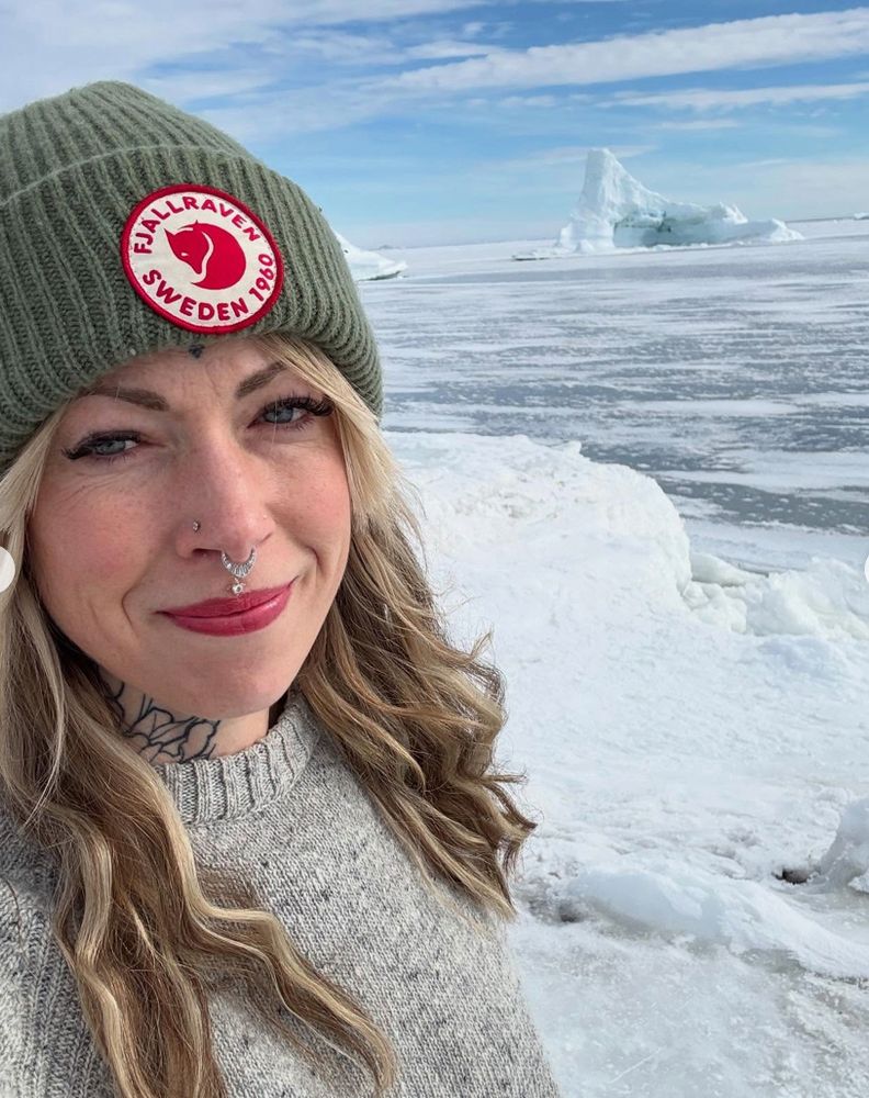 selfie pic of Helen Mort, a white woman with long blonde hair and a nose piercing, in a green hat, smiling into the camera, with a background of icebergs and frozen sea under a blue sky.