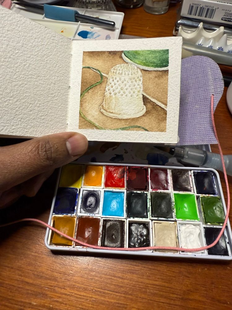 Photo of a hand holding a small watercolor notebook with a painting of a thimble and green spool of thread over a wooden surface with a watercolor palette and water brush on it. 