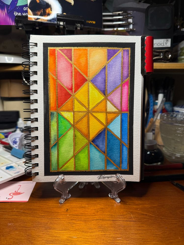 Photo of a stained glass pattern watercolor painting in a watercolor notebook displayed on a clear plastic easel atop a wooden desk with clutter in background. 