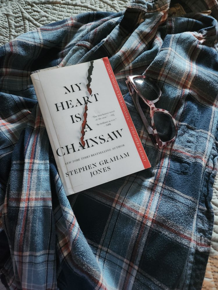 My Heart is a Chainsaw by Stephen Graham Jones paperback book and a pair of sunglasses on a plaid flannel shirt.