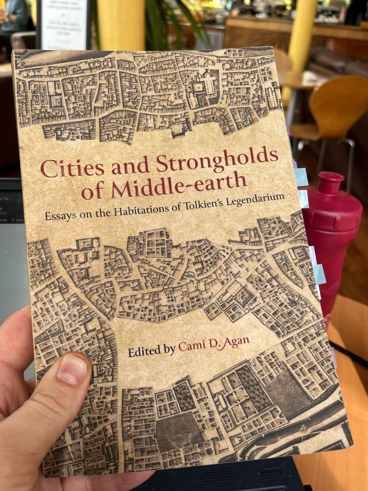 The book Cities and Strongholds of Middle-earth: Essays on the Habitations of Tolkien's Legendarium, edited by Cami D. Agan. The cover depicts a city grid viewed from overhead; in the background of the photo is a coffee shop full of tables, couches, and a decorative plant.