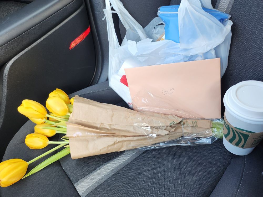 A picture of the care package I brought my mom. On a car seat are a bouquet of yellow tulips, a Starbucks hot drink cup, a peach colored card with the word Mom on it, and a plastic bag with medicine in it for my mom.
