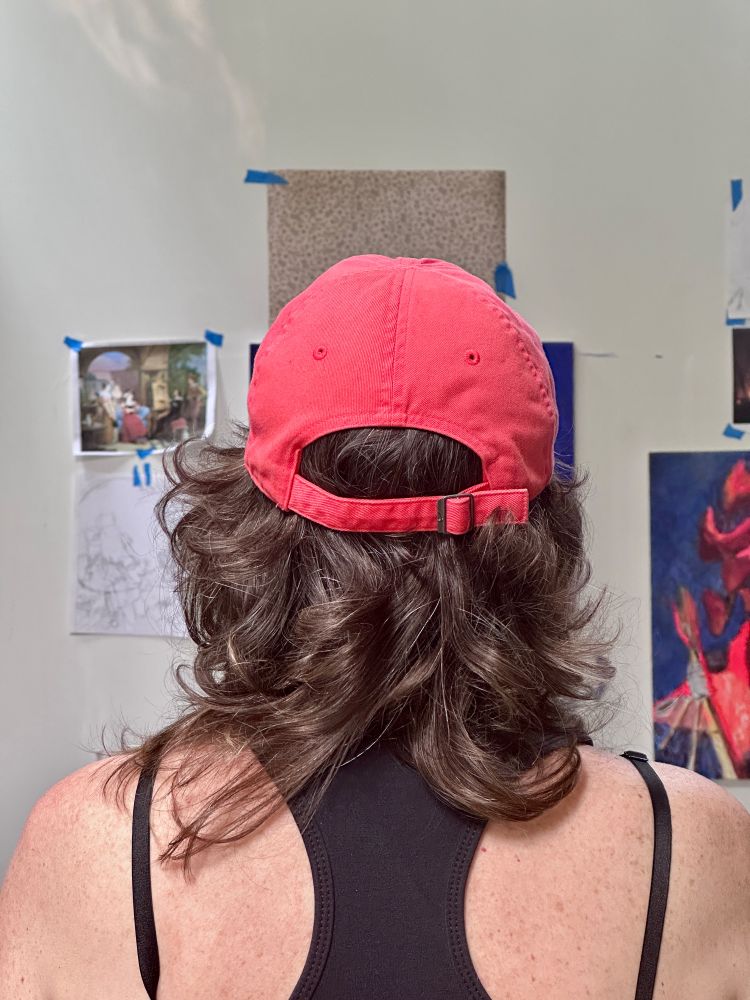 Back of a person’s head with long curly grey-brown hair and black tank top wearing a pink baseball cap standing in art studio