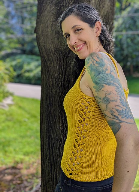 A woman standing in front of a tree. She's wearing a hand knit yellow tank top with a butterfly lace pattern running down the side. She's got blue tattoos from shoulder to elbow and is looking over her shoulder and smiling. 