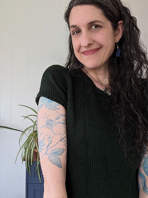 A woman standing in front of a while wall and smiling. She's wearing a hand knit, dark green tee and blue sea glass earrings. She has long black hair streaked with gray and blue tattoos on both arms. 