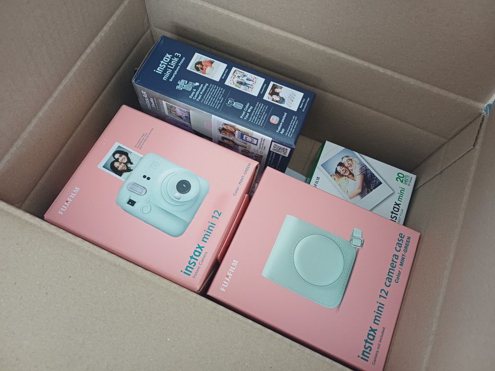 A photo of the contents of a cardboard box. Inside are more boxes, two salmon pink ones containing an instant camera and a camera case, a navy box with a small printer, and a white box with instant film for the devices.