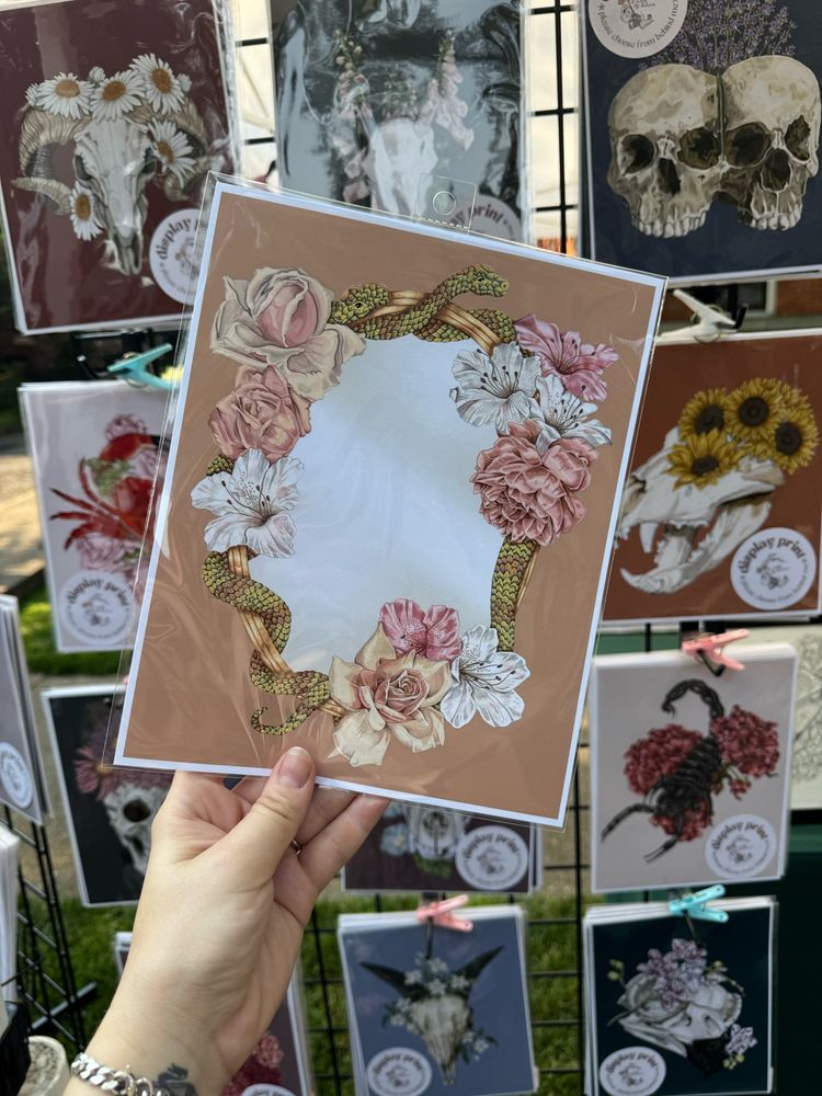 A hand holding a color drawing of snakes and roses around a mirror. Other art is hung in the background.