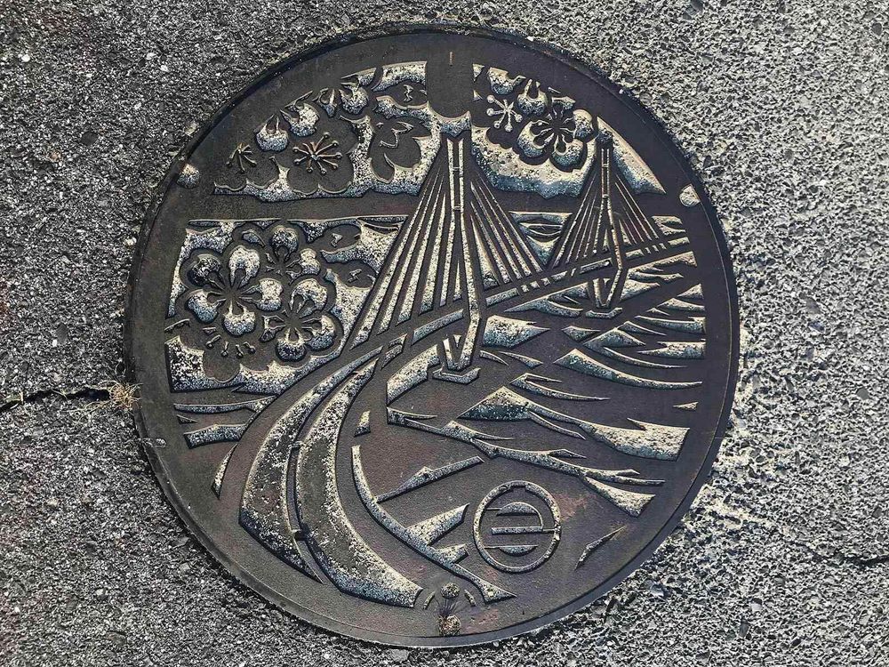 Ornate metal manhole cover depicting a bridge over water, with floral designs.