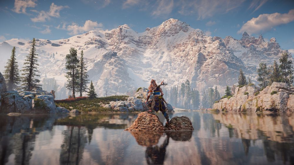 A scenic view of a mountain range, a crystal clear lake, and Aloy standing on a small wall in the middle of the lake looking towards the mountains.