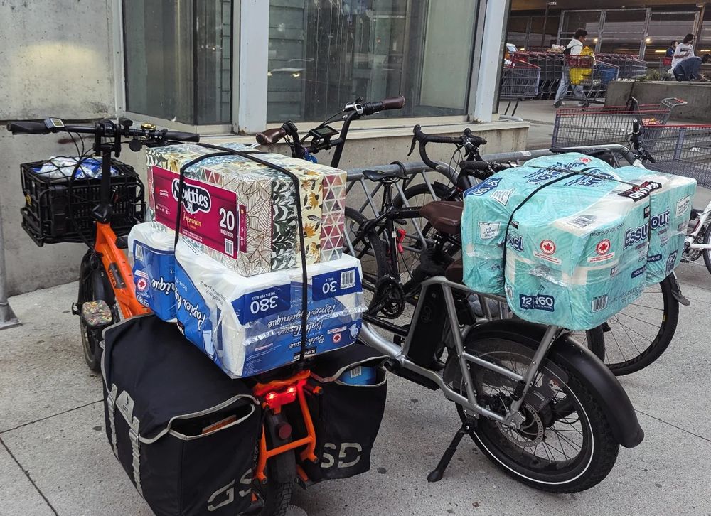 Two ebikes are loaded with groceries & large packs of toilet paper, Kleenex, & paper towel outside a Costco