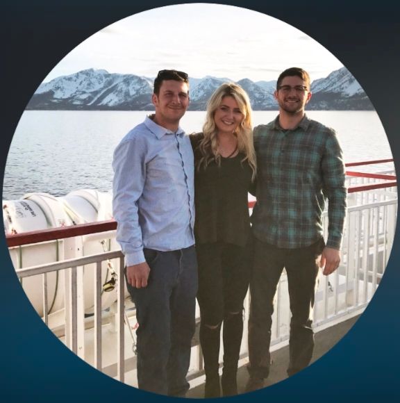 Two white men & a white woman standing on the outdoor deck of a shop, water & mountains in the background 