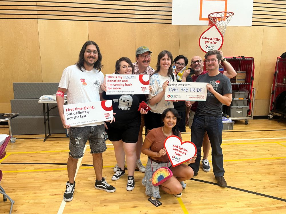 I group of people is crowded together in what looks to be an elementary school gym. They are all smiling and holding various signs from The Canadian Blood Services, including one that is written on that says "Saving lives with CAG 938 Pride!"