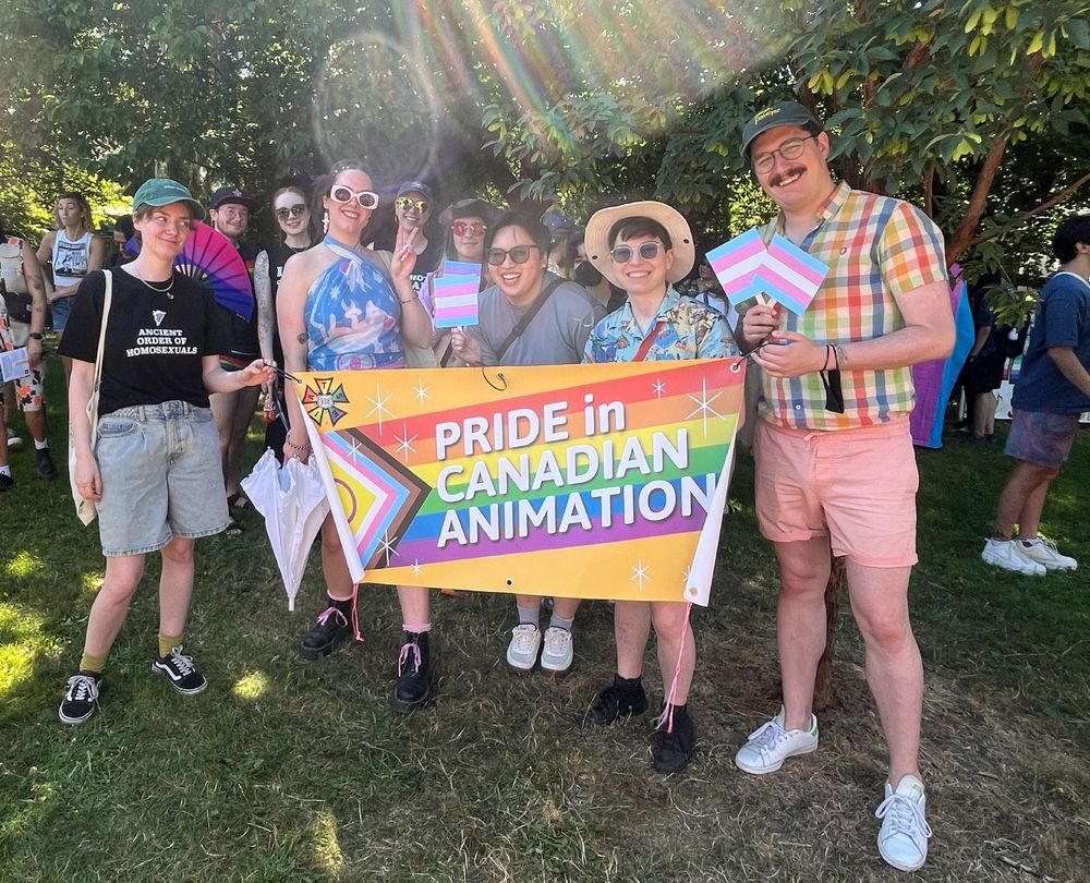 A different photo of a different group of happy people in a different grassy field. Several of the people are holding little trans flags and two of the people are holding up a banner that has the colours of the Progress Pride flag that reads "PRIDE in CANADIAN ANIMATION"