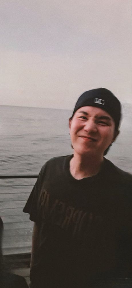 A slightly blurry pic of Yoongi. He’s scrunching his nose cutely, wearing a black t-shirt and black hat. The ocean is in the background.