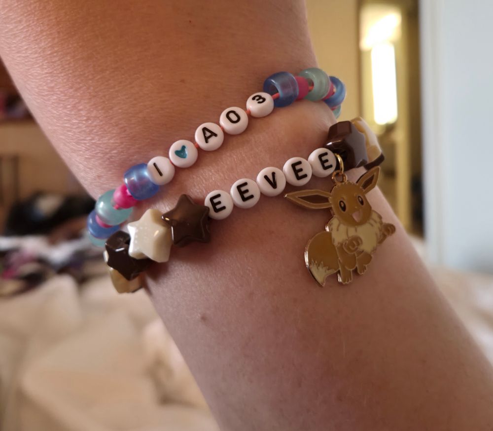 Two friendship bracelets one labeled EEVEE with a charm of the Pokémon and the other is labeled I Heart Symbol AO3.