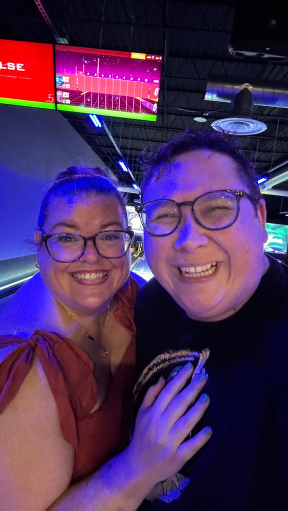 A selfie of Jackie and Jess at a bowling alley, bathed in a weird blue tint from a blacklight. 