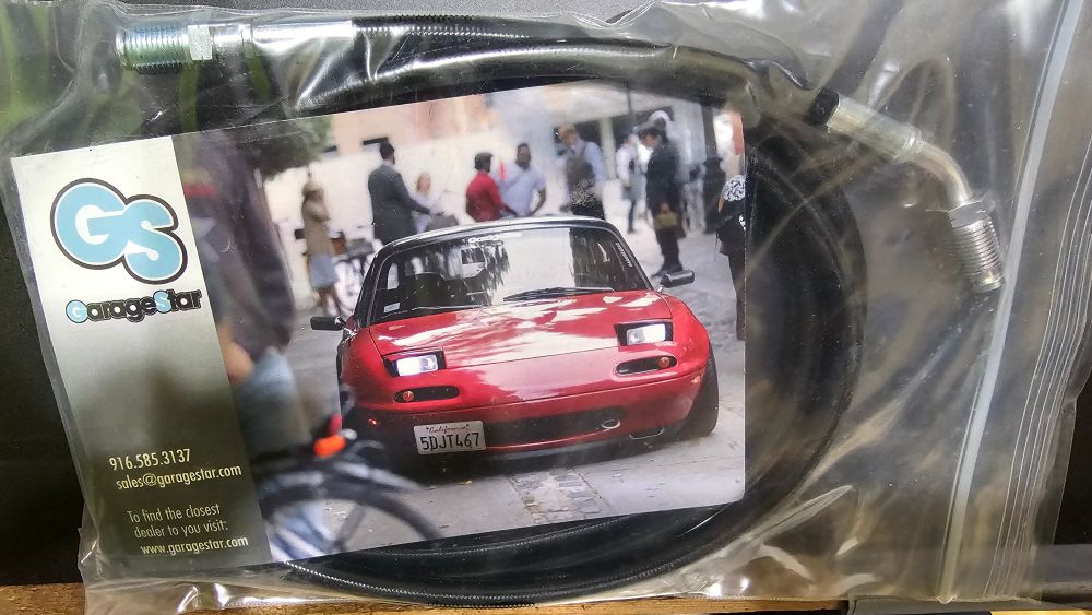A clear ziploc bag with a black, one-piece hydraulic clutch line inside aling with a business card that has a picture of a red Mazda Miata and the company's information. The company is GarageStar.