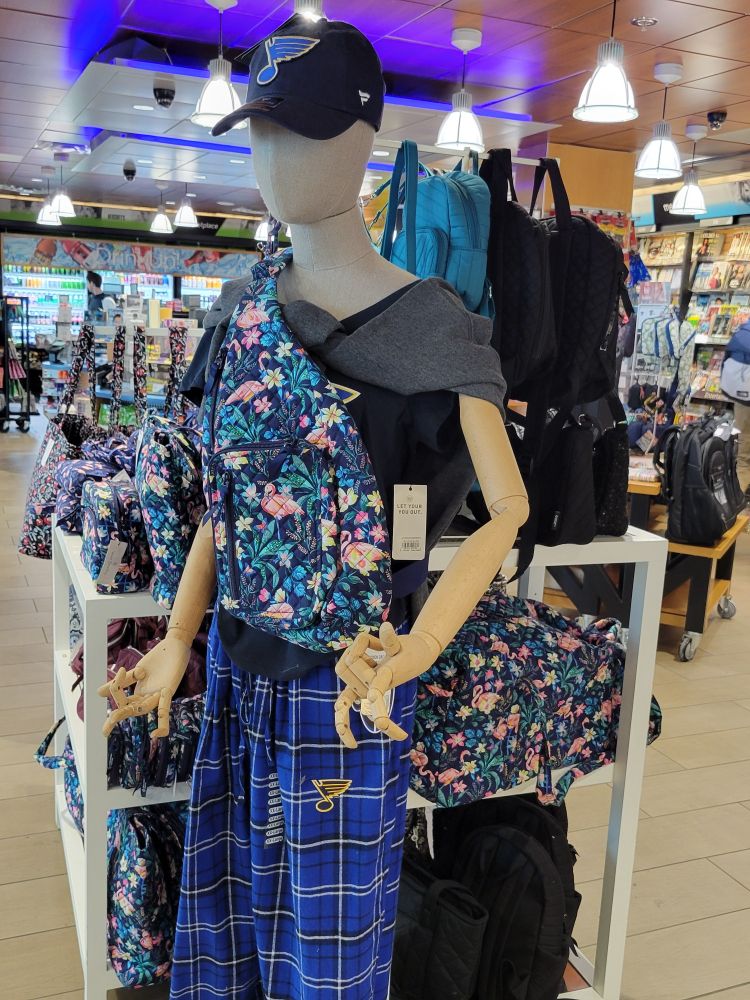 A mannequin in a store in the St. Louis airport. It is wearing a floral bag, a hat with a blue music note on it, and plaid pants with a similar looking note on it. The fingers are in angles that no person could naturally put them.