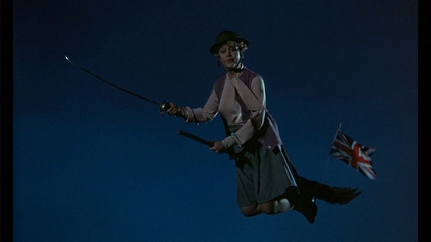 Angela Lansbury as the witch Eglantine Price in Bedknobs and Broomsticks. She is flying on a broom with a small Union Jack flag stuck in the bristles, and she is holding a sword aloft in her right hand. 