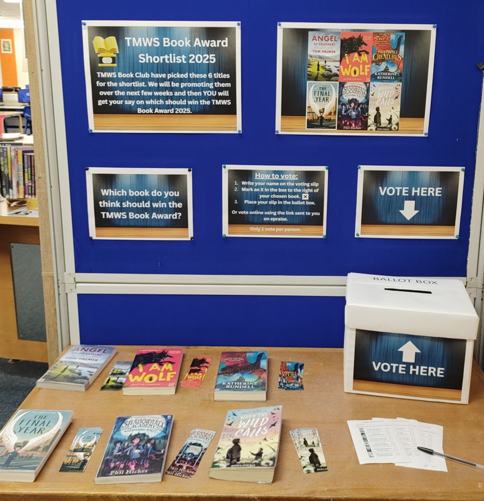 A blue display board shows the front covers of the shortlisted books and instructions on how to vote. In front is a wooden table with a ballot box, voting slips, and a copy of each of the books:
Angel of Grasmere by Tom Palmer
I Am Wolf by Alastair Chisholm
Impossible Creatures by Katherine Rundell
The Final Year by Matt Goodfellow
Shadowhall Academy: The Whispering Walls by Phil Hickes
When The Wild Calls by Nicola Penfold