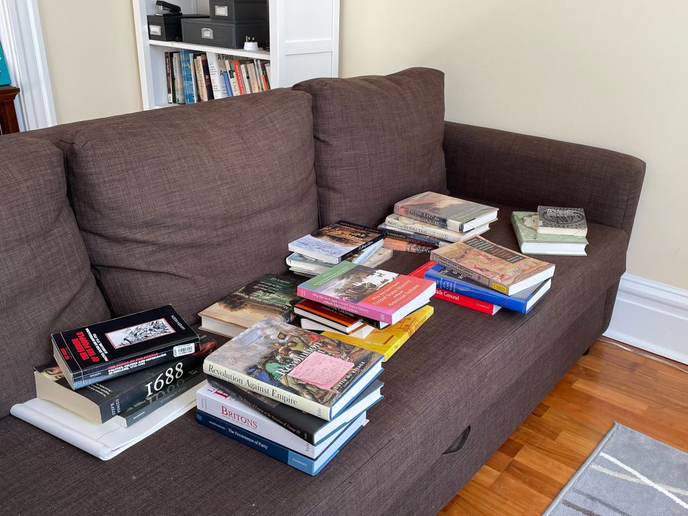 Many books stacked haphazardly on a sofa. 
