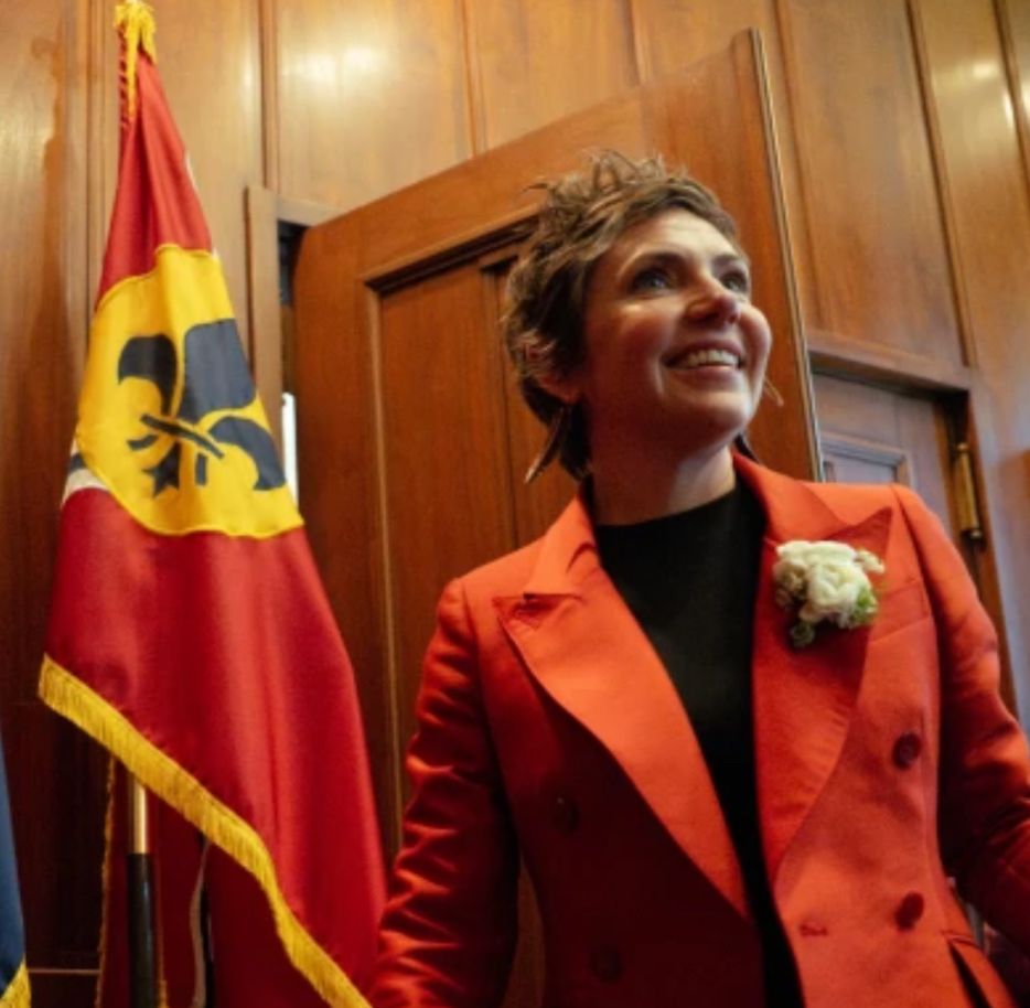 St. Louis mayor Cara Spencer, a 40-something woman with a pixie haircut, wearing a blazer with wide lapels and a huge boutonnière in front of a flag and wood-paneled walls. 