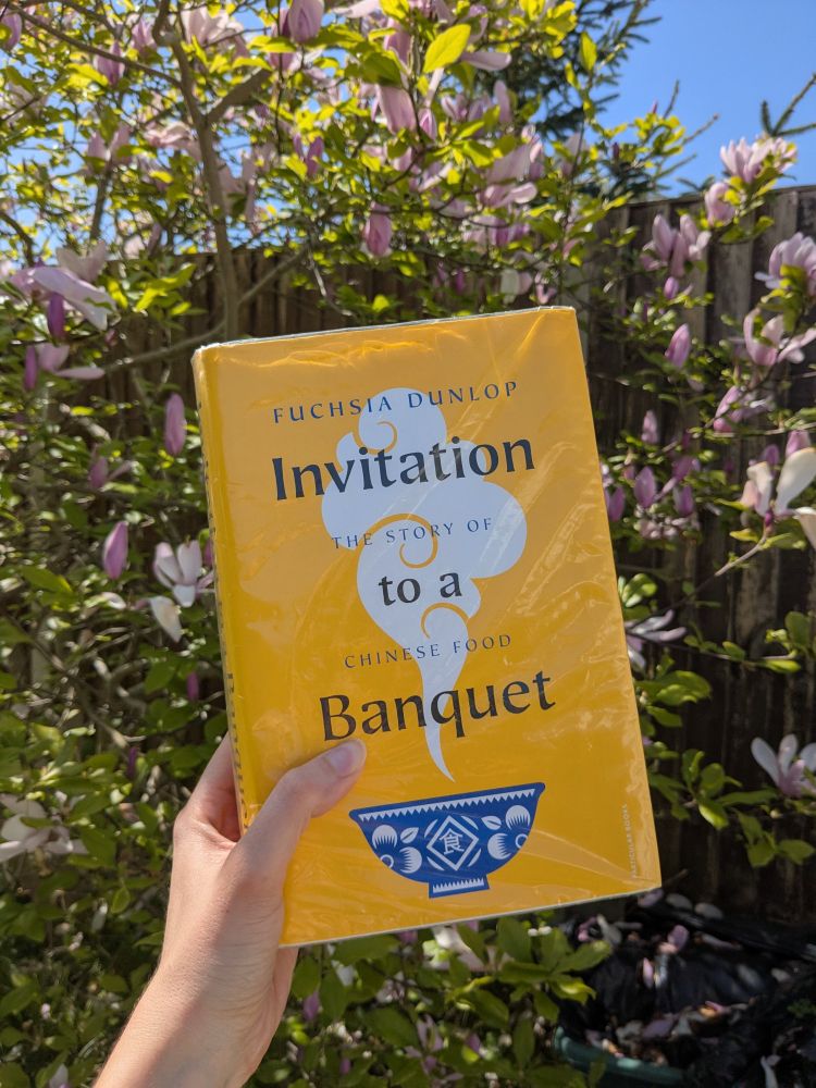 A hand holds a hardback copy of "Invitation to a banquet; The history of Chinese food" by Fuchsia Dunlop in front of a blooming magnolia tree. The book has a bright yellow cover with a drawing of a white and blue porcelain bowl from which a white cloud of steam arises. Published by Particular Books.