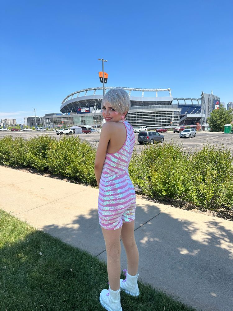 me (a mid-20s woman with a short, blonde pixie cut wearing a handmade pink and iridescent striped bodysuit), standing on a side walk in front of Mile High Stadium in Denver, hours before the Eras Tour concert on 7/15/23