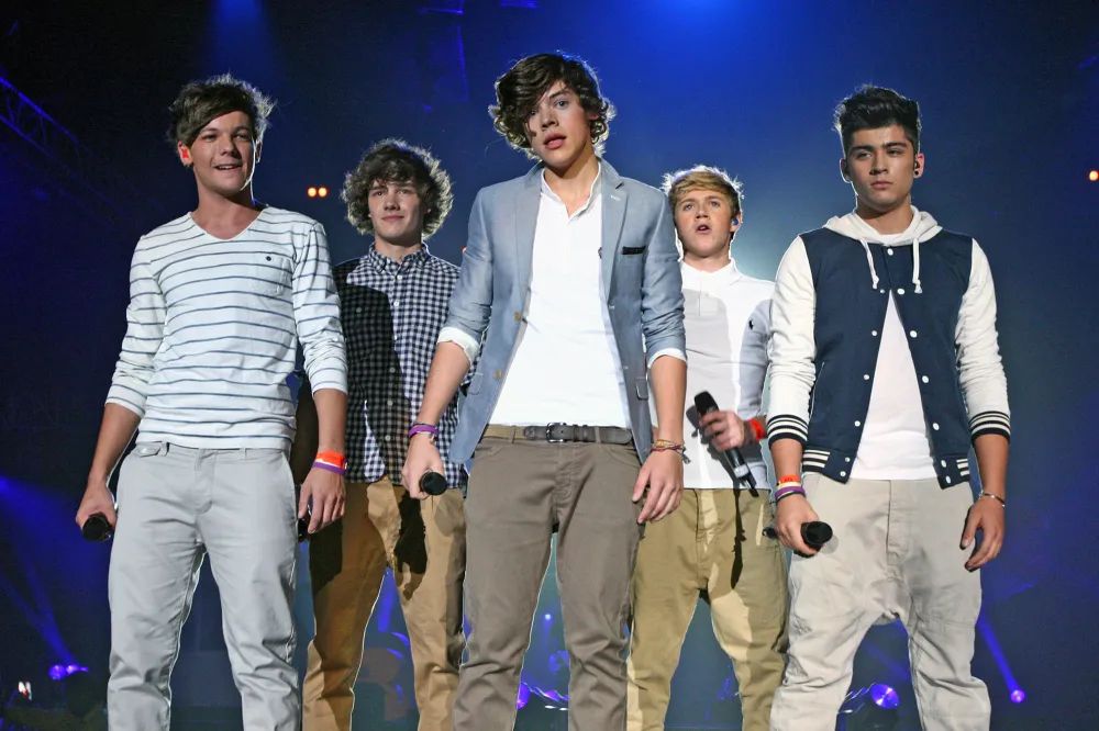 One Direction performing on British talent TV show ‘X Factor’ c. 2010. In order from left to right: Louis Tomlinson (white and black horizontal striped shirt and gray khakis), Liam Payne (black and white checkered shirt and tan khakis), Harry Styles (white shirt, silver-blue blazer, light brown khakis), Niall Horan (white polo, tan khakis), and Zayn Malik (white shirt, navy and white varsity style jacket, saggy beige khakis). All are holding microphones in their hands and are performing on a stage surrounded by blue lighting