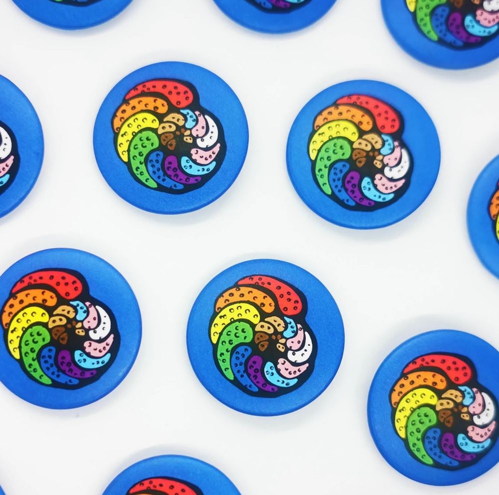 Button pins from the Etsy store of the queer art collective, Palaeoiris, featuring a foraminifera test (or shell) in the colors of the progress pride flag. The background of each pin is solid blue. Buttons are equally spaced apart from one another on a white background, depicting full or partial images of twelve pins.