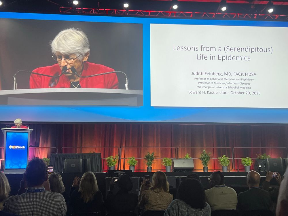 A picture of the IDSA stage featuring Judith Feinberg at the lectern and on the big screen with her lecture titles, ‘Lessons from a (Serendipitous) Life in Epidemics’, a talk about her career treating HIV, HCV, and substance use disorder; preventing the transmission of infectious diseases; and fighting stigma.