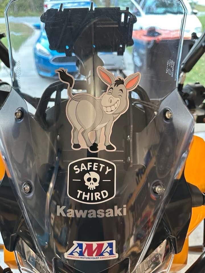 The windscreen of a motorcycle. It has vinyl stickers on it. One is a donkey looking back coyly over its shoulder. One says “Safety Third” and the last says AMA, short for “American Motorcyclist Association”. 