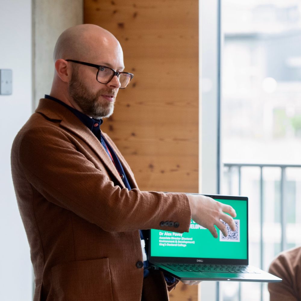 A photograph of a bald and bearded man with glasses. He is holding a laptop and gesturing at the screen while delivering a talk. 
