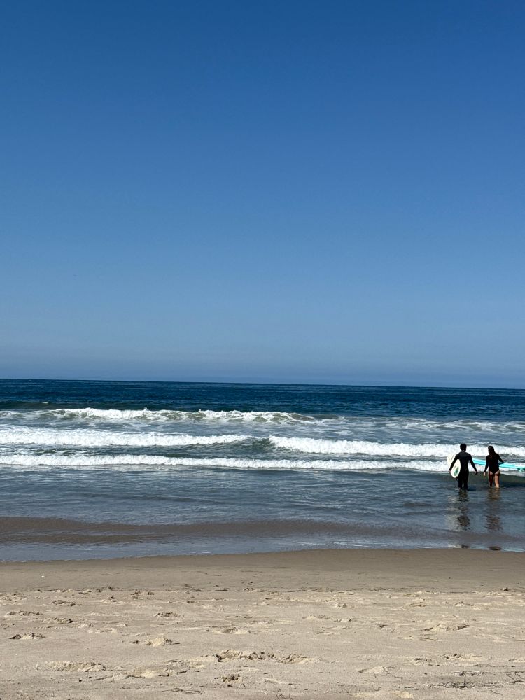 Small waves are breaking on the beach. A pair of surfers are walking out. Not a cloud in the sky, this is probably what Heaven looks like if I were to guess.