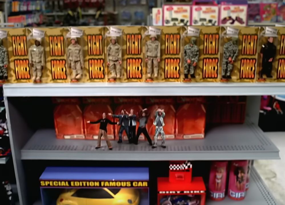 a screenshot from nsync's "it's gonna be me" music video, portraying the band members as plastic dolls in a toy store surrounded by fictional non-copyright-infringing toys labeled "fight force," "special edition famous car," "dirt bike," and "girl"