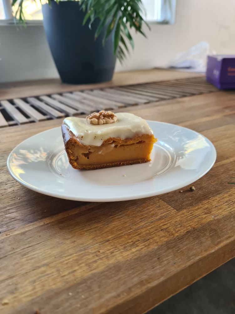 A slice of honey pumpkin cake with maple cream cheese icing on a white plate, placed on a wooden table. Fronds from a parlour palm, and a children's game can be seen in the background.