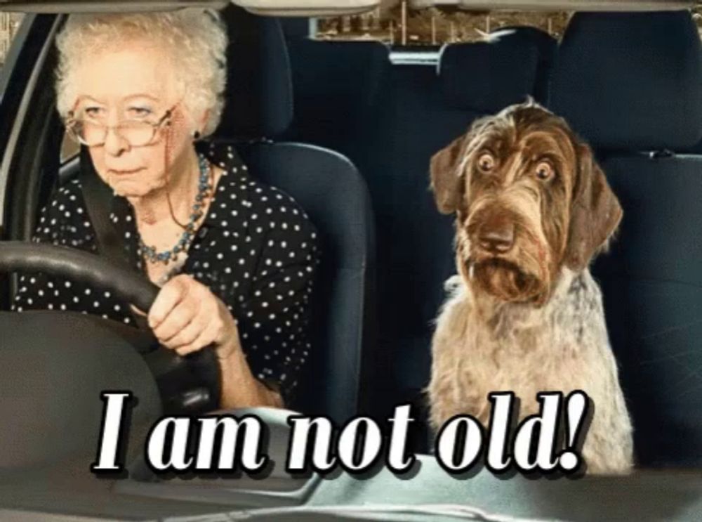 an elderly woman is driving a car with a dog in the back seat and the caption i am not old