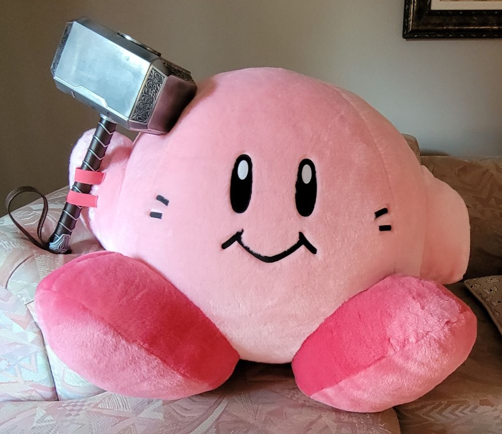 A giant Kirby plush sitting on a couch wielding Thor's mythical hammer Mjolnir.