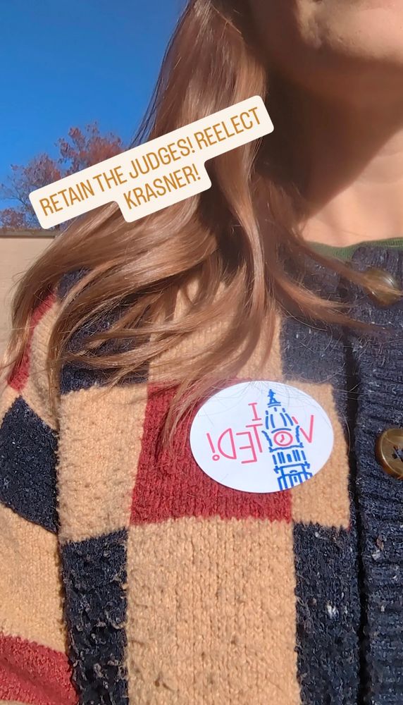 photo of my shoulder and hair. blue brown and red checked cardigan with an I VOTED sticker. the sky is blue. text reads RETAIN THE JUDGES! REELECT KRASNER!
