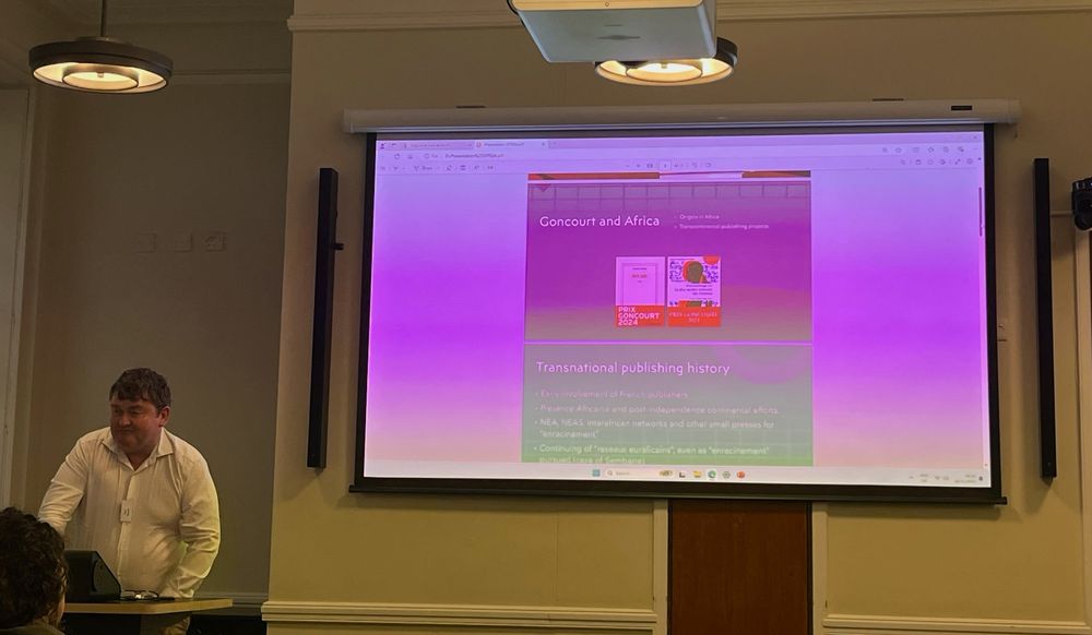 Presenter standing in front of a lectern, with a power point displaying pictures of two books