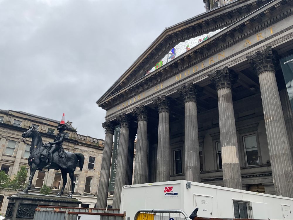 On the left, is a statute of a man (The Duke of Wellington) on horse back, with a cone on traffic cone on its head. On the right is the Musuem of Modern art, a building with columns in front of it. There is a white rectangular container in the foreground. 