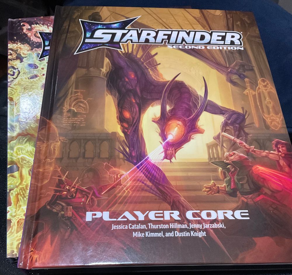 Most of the image is filled by a boom, Starfinder Second Edition Player Core, with another similarly coloured book partially visible on the left hand side, against a dark and blue background. 