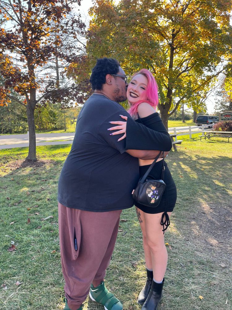 a large black man embraces his pink haired white girlfriend who is beaming while leaning into his embrace. they are so in love, and happy to be together.