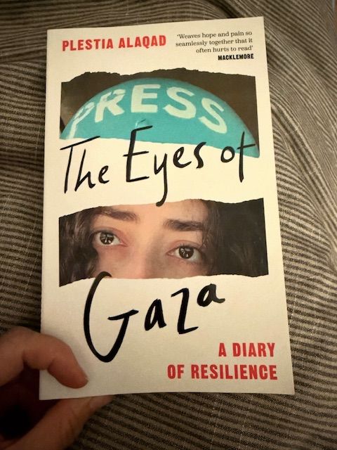 Hand holding a book featuring a turn image of Plestia's face - you can see her eyes and press helmet - with the type: PLESTIA ALAQAD, THE EYES OF GAZA: A DIARY OF RESILIENCE
