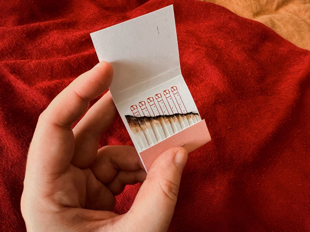 photo of me holding a matchbook, open this time. the interior is white and shows matches burned down in a diagonal line. behind them is revealed an illustration of wolfwood's vials in red. my hope with this is that it'd give a sense of time running out, and evoke a parallel use of wolfwood's two vices of choice (cigarettes and serum)