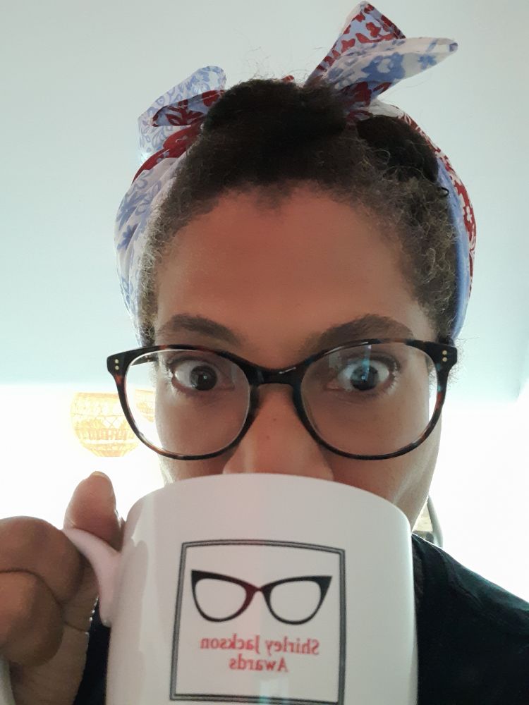 Pic of author Eden Royce  with her morning coffee. She is wearing eyeglasses with dark tortoiseshell frames and her hair is tied up in a white bandanna with read and blur flowers. She is looking at the camera and sipping out of a white coffee mug with the iconic Shirley Jackson Awards logo: black cat-eye glasses over the red lettering.