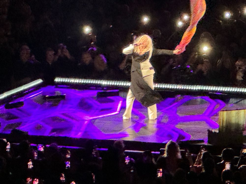 Cyndi Lauper on a lighted platform holding a flowing rainbow scarf. She’s singing “True Colors”.